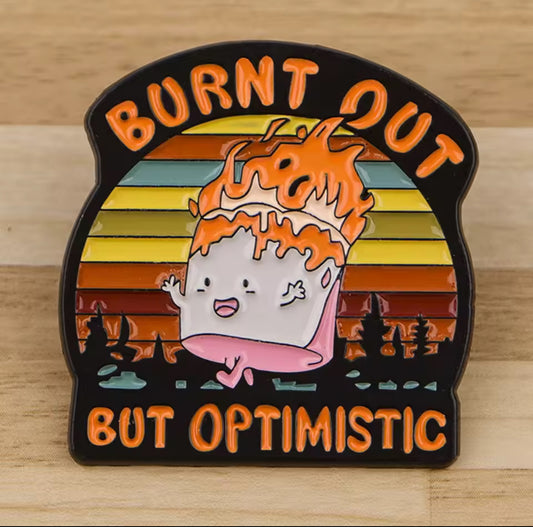 “Burnt Out But Optimistic enamel pin featuring a smiling cartoon character on fire with retro rainbow background and bold orange text.”