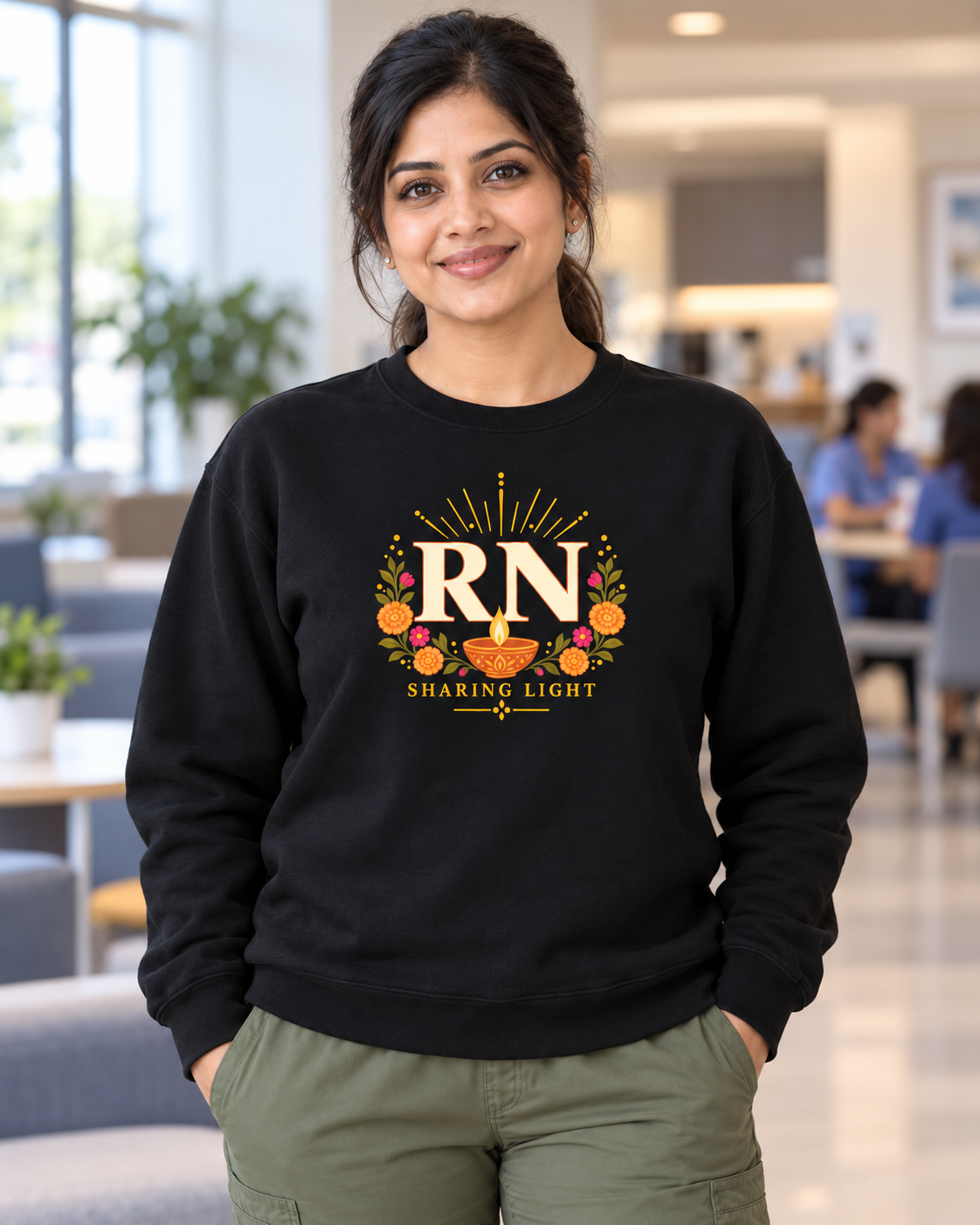 Female nurse wearing a black Nursify RN Diwali crewneck sweatshirt with ivory RN lettering, a diya lamp and colourful flowers, paired with olive scrub pants.