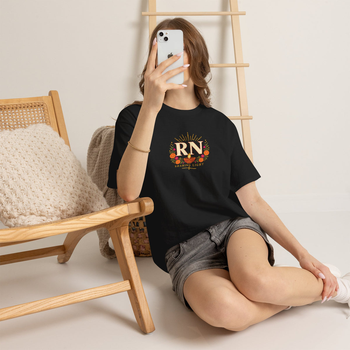 Woman wearing a black Nursify RN Diwali T shirt with a centred diya and floral design, seated beside a chair in grey shorts.