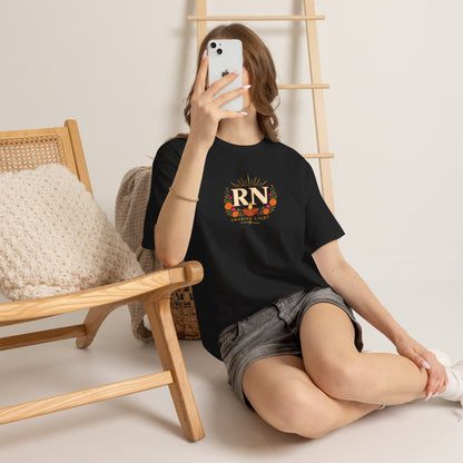 Woman wearing a black Nursify RN Diwali T shirt with a centred diya and floral design, seated beside a chair in grey shorts.