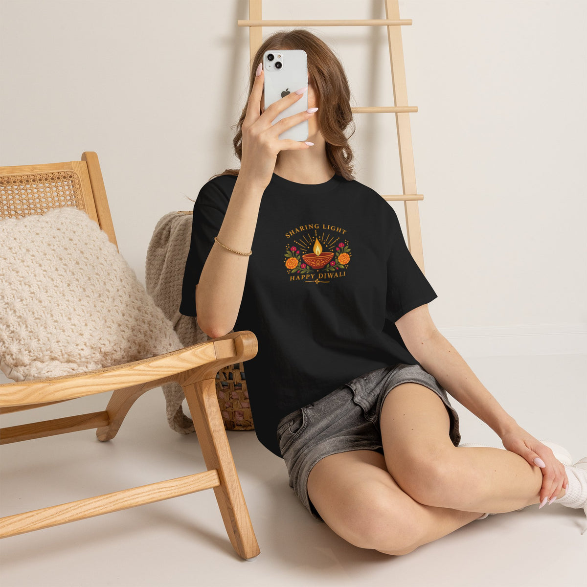 Woman wearing a black Sharing Light Happy Diwali T shirt with a diya and floral print, seated beside a chair in grey shorts.