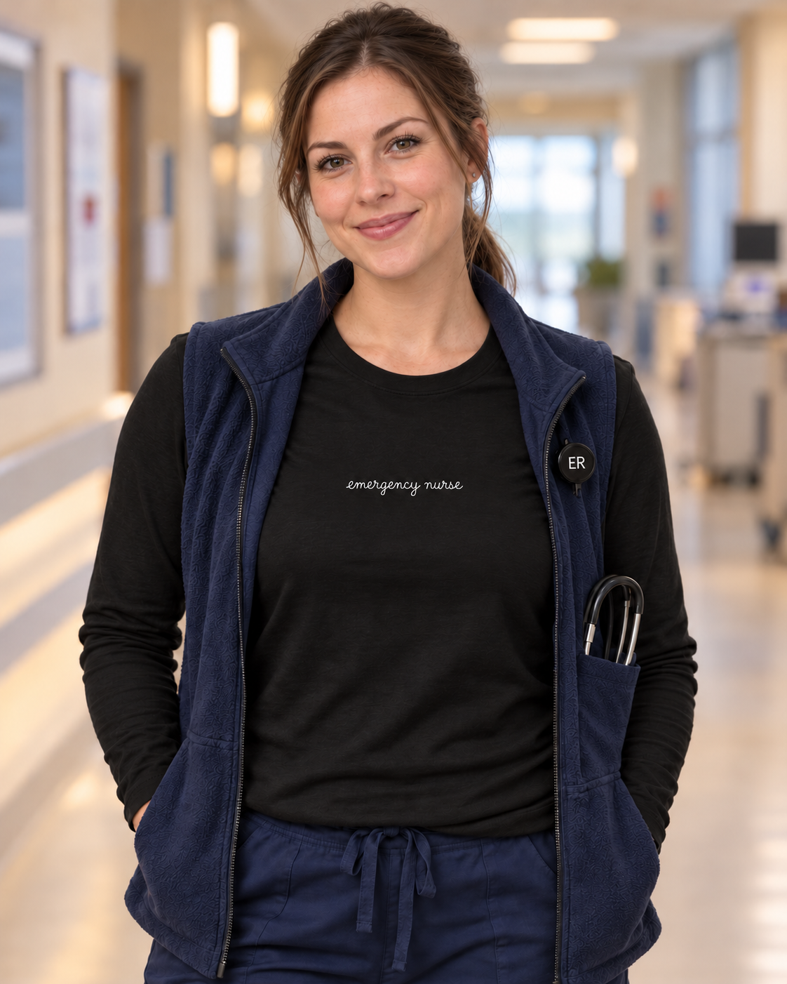 Young nurse wearing a black Emergency Nurse script long sleeve shirt under an open navy vest, with the centred white lettering visible.