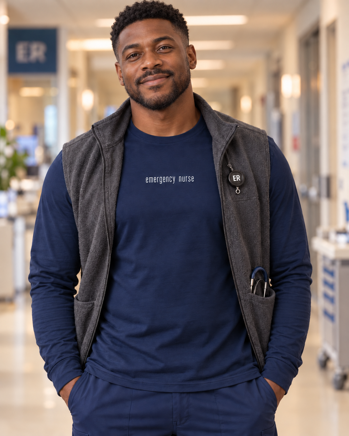 Male nurse wearing a navy Emergency Nurse long sleeve shirt under an open charcoal vest, with the centred white lettering visible.