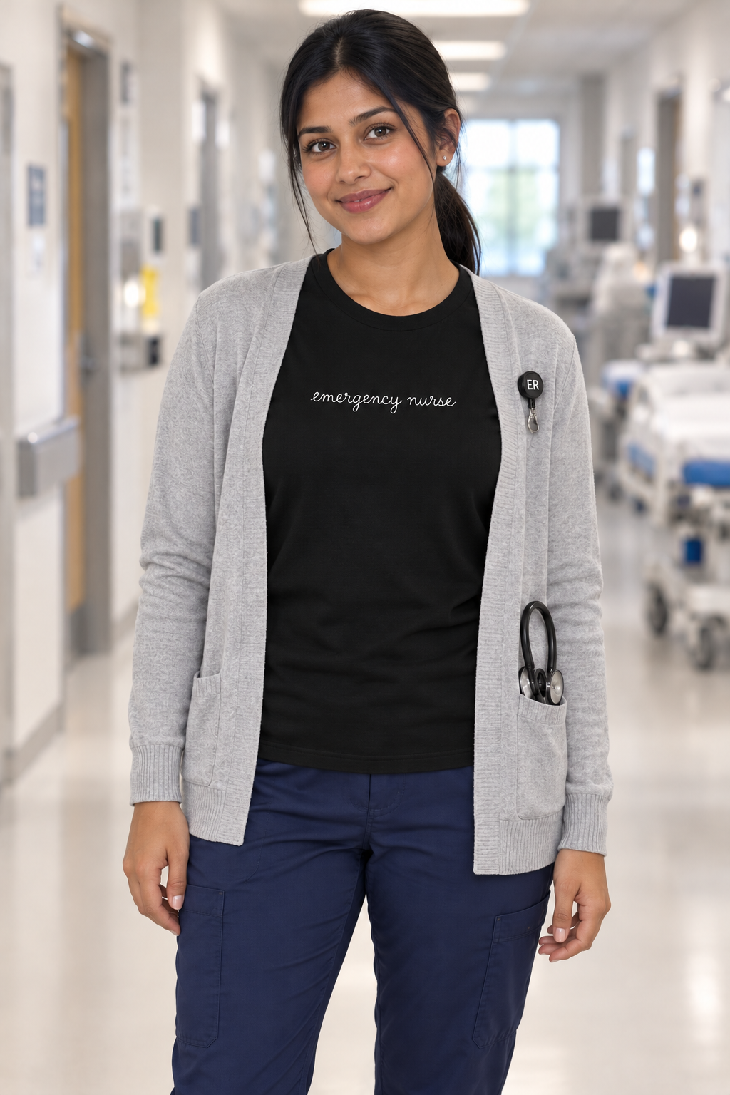 Young East Indian nurse wearing a black Emergency Nurse script shirt under a grey cardigan, open to show the centred white lettering.