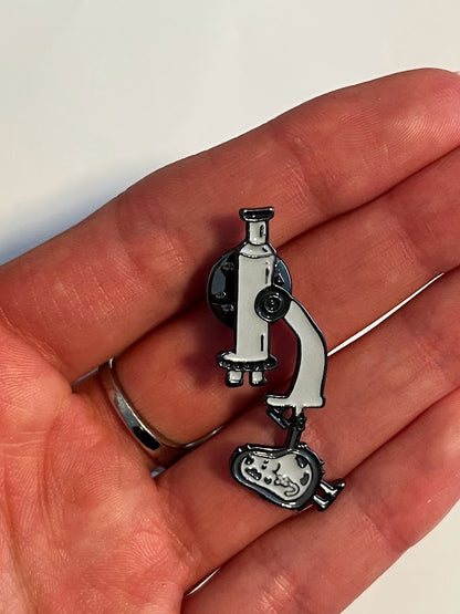 Microscope enamel pin in grey with smiling cell detail | 1.7 cm x 4.2 cm compared to a female average sized hand 