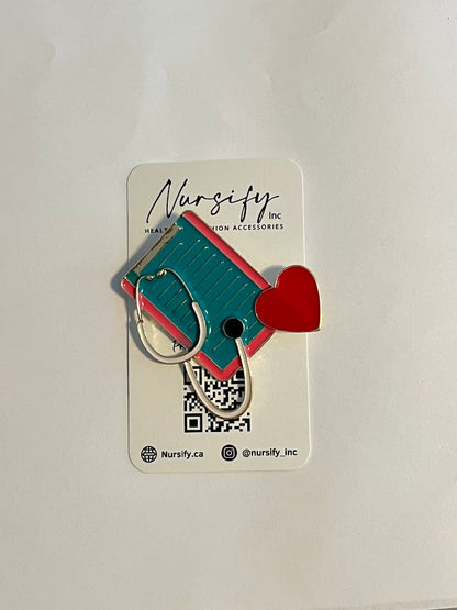 Clipboard stethoscope enamel pin with white and gold stethoscope, pink and teal clipboard, and red heart | 4.8 cm x 5 cm size compared to packaging