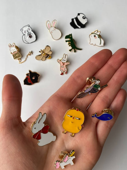 this images shows a hand holding an Assortment of small animal and creature enamel pins in different illustrated designs, shown together on a white background to display selection and scale. The individual hand hold a bunny, unicorn, whale, baby chick, and a bunny on a witch broomstick  