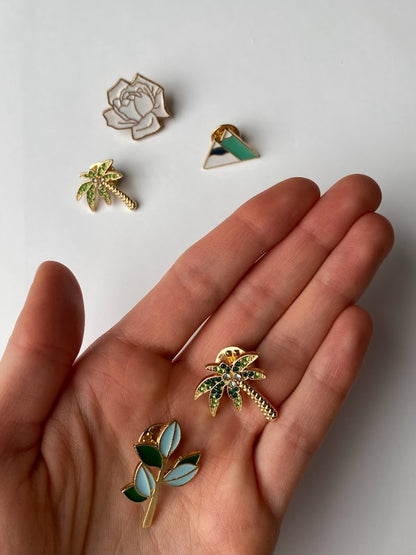 a hand holding a photo of five pins which include a white rose with gold accents a dark green palm tree lighting palm tree mountains and a branch with blue and green leaves. 