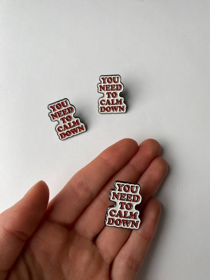 You need to calm down enamel pin in orange and white text design | size shown compared to an average-sized woman’s hand