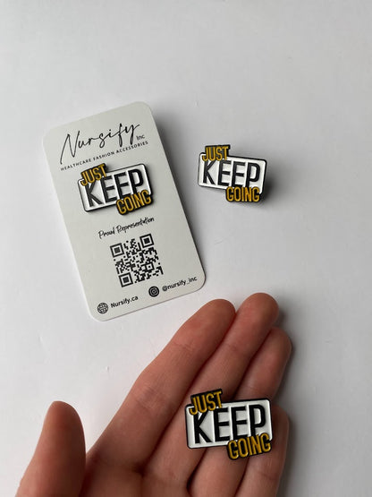 Just keep going enamel pin in white, black, and yellow text design | size shown compared to an average-sized woman’s hand