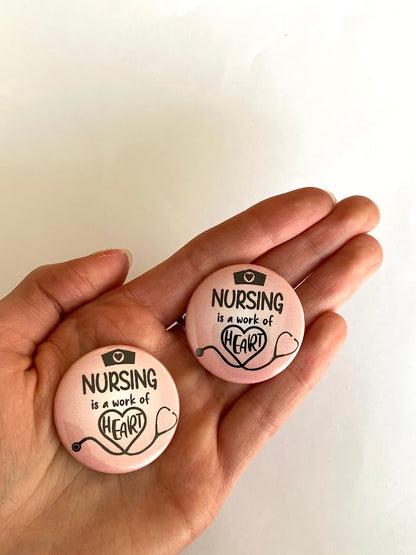 Two pink Nursing Is a Work of Heart button pins shown in a hand for scale