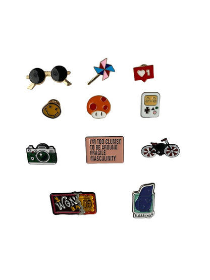 this picture includes an assortment of pins  which are Sunglasses, a Pinwheel, Like Button, Smiley Face, an Orange Mushroom, Game Boy, Green Camera, Fragile Masculinity Quote, Bicycle, Wonka Chocolate Bar and Avogadro Constant and Mole.