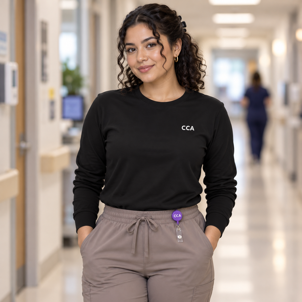 Healthcare worker wearing a black Nursify CCA long sleeve shirt with taupe scrub pants and a purple CCA badge reel.