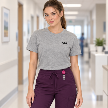 Healthcare worker wearing a heather grey Nursify CTA T-shirt tucked into plum scrub pants with a small pink CTA badge reel.