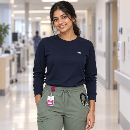 Nurse wearing a navy Nursify RN long sleeve shirt tucked into sage scrub pants, with a pink badge reel at the waistband.